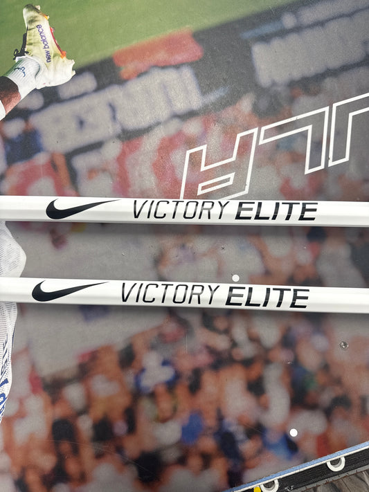 Two white Nike Victory Elite Women’s Lacrosse Shafts, each with black text and the Nike Swoosh, rest on a blurred sports field while part of a player’s shoe is visible in the upper left.