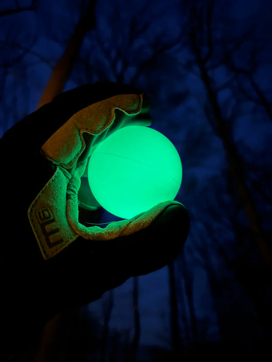 A gloved hand holds a glowing Firefly Glow in the Dark Lacrosse Ball against a dark blue night sky with silhouettes of leafless trees. Shown: Firefly 3 pack with black light flashlight.