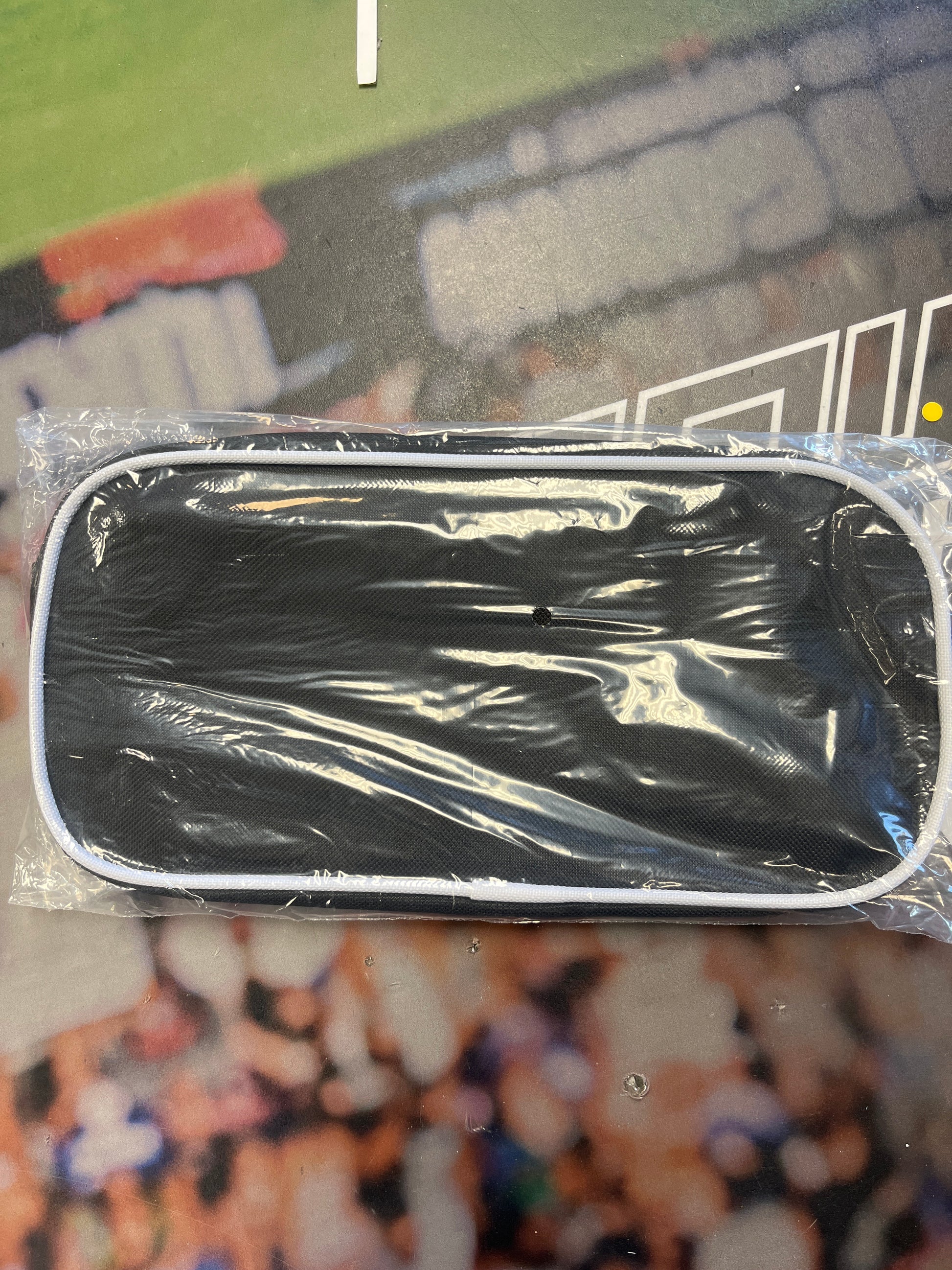 The Sanderson Lacrosse Howies accessory bag is black, rectangular with rounded corners, and wrapped in clear plastic. It features a white border and a small hole in the center, resting on a colorful, blurred surface.
