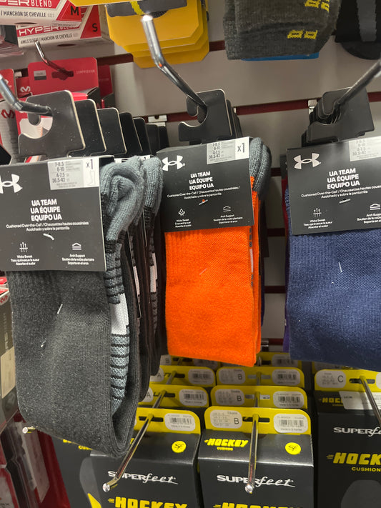 Under Armour Team Cushioned Socks in gray, orange, and blue hang on display hooks above boxes of Superfeet insoles. These crew socks offer comfort and moisture-wicking performance for athletes.