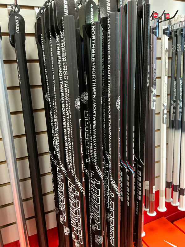 Several Loading Northmen Lacrosse Shafts by Sanderson Lacrosse, black with “NORTHMEN” vertically printed, are displayed upright on a rack alongside other sticks in a sports equipment store.