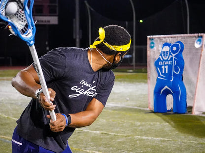 Wearing a yellow bandana and black t-shirt, a person prepares for realistic practice by aiming a lacrosse ball at the Elevate 11th Man Goalie Pro by Elevate on a sports field at night.