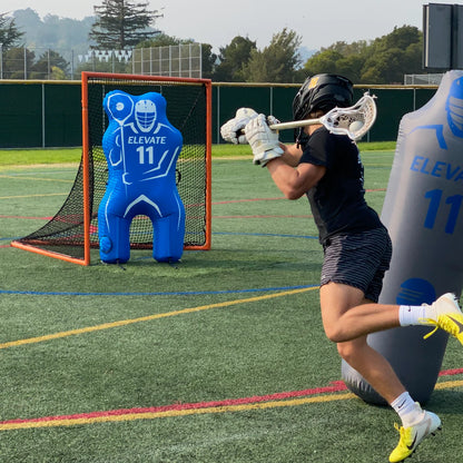A lacrosse player in black and stripes practices shooting at an Elevate 11th Man Goalie Pro by Elevate. Another inflatable dummy stands nearby on the turf, with trees and a fence in the background.