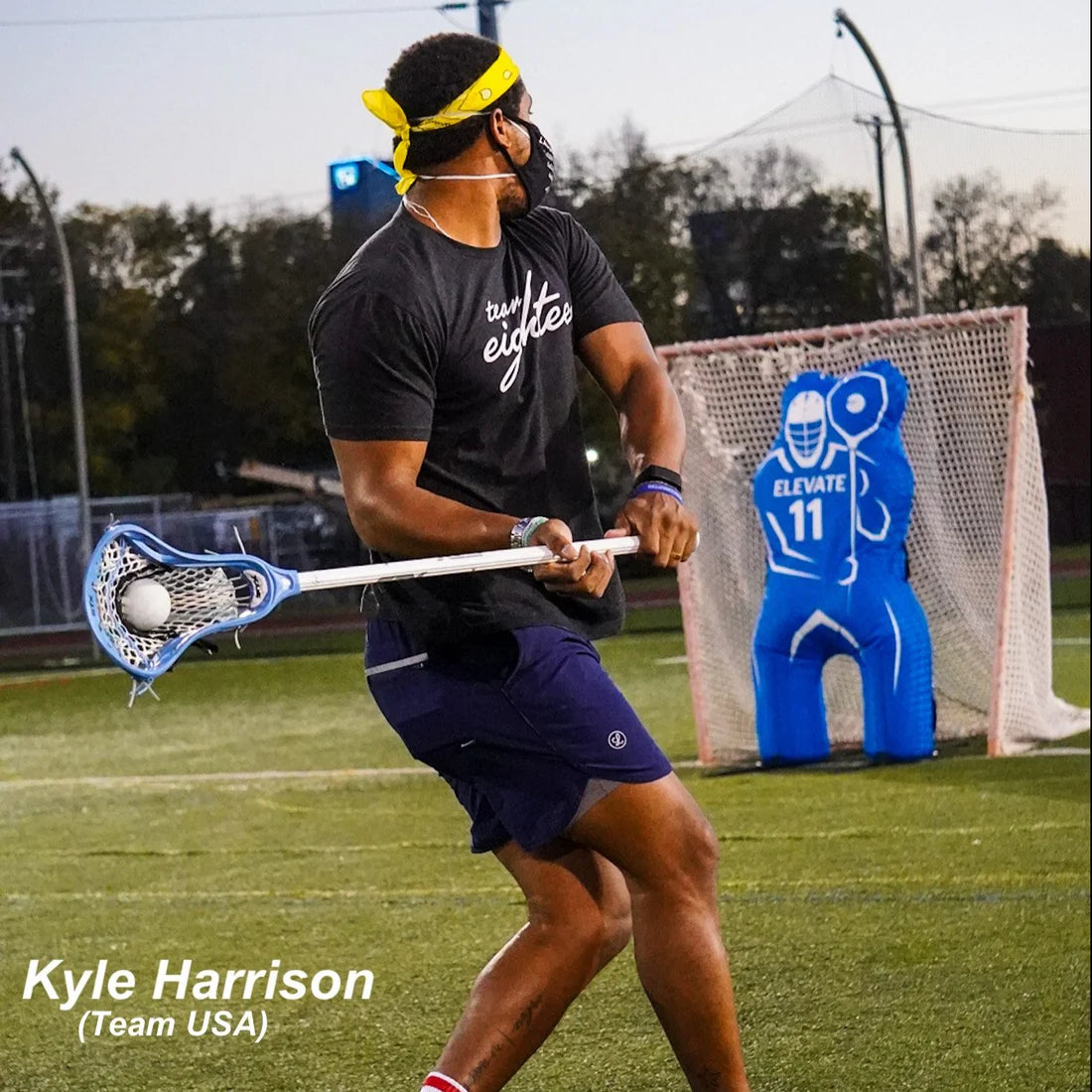 A man in a yellow headband and black mask practices lacrosse goalie moves on a field, swinging his stick near the Elevate 11th Man Goalie Pro by Elevate. Text: Kyle Harrison (Team USA).