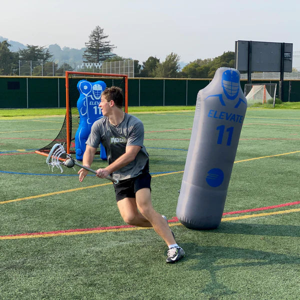 A young man in athletic gear dodges around an Elevate 11th Man Goalie Pro by Elevate on an outdoor turf field. Another dummy and a lacrosse net appear in the background, ideal for lacrosse goalie training. Trees and a scoreboard stand beyond the fence.