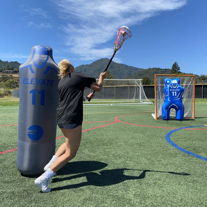 A lacrosse player practices shooting outdoors, aiming at a goal with a blue target and Elevate’s Elevate 11th Man Goalie Pro inflatable goalie dummy, ideal for realistic lacrosse goalie training. Soccer goals and mountains are visible in the background.