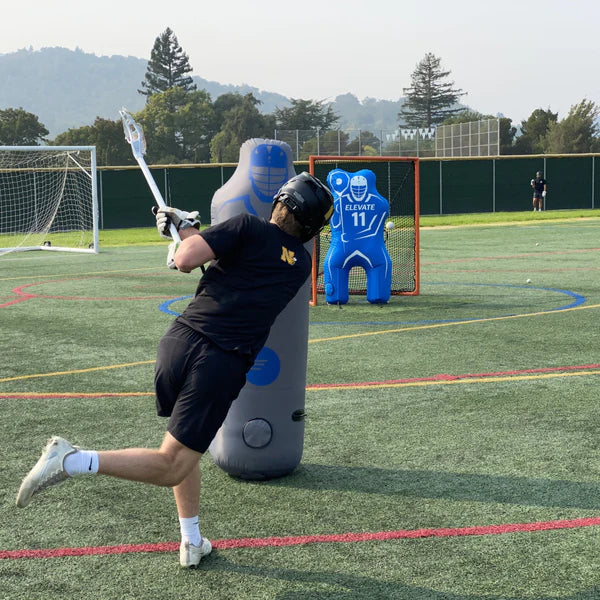 A lacrosse player in black practices shooting on outdoor turf, aiming at the Elevate 11th Man Goalie Pro by Elevate, an inflatable goalie in front of the net, with trees and hills visible in the background.