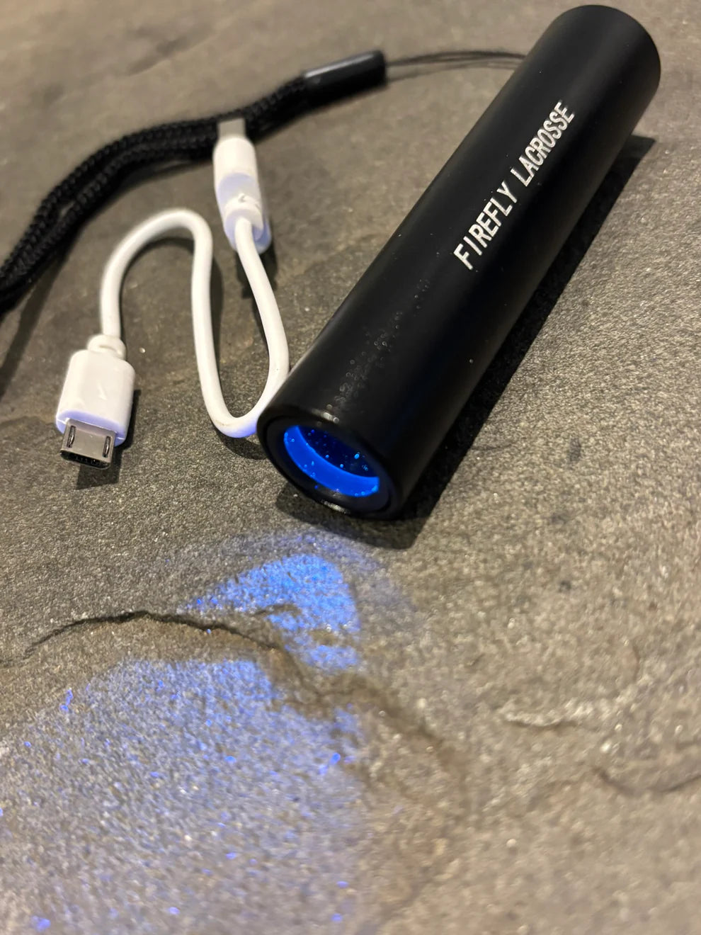 The Firefly Glow in the Dark Lacrosse Balls 3-pack comes with a black cylindrical FIREFLY flashlight emitting blue light to charge the balls. A white micro USB charging cable is included for easy recharging.