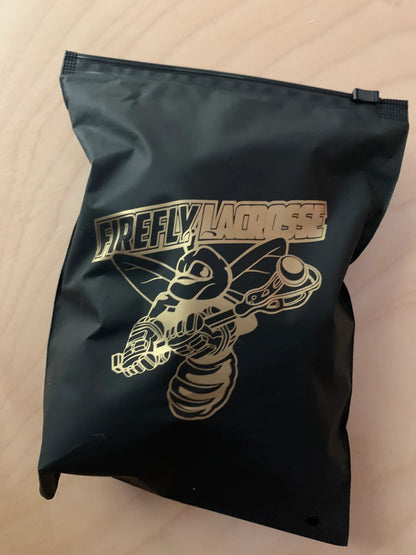 A black pack with a gold robotic insect and "Firefly Lacrosse" text sits on a light wood surface, featuring Firefly Glow in the Dark Lacrosse Balls (3-pack) with black light flashlight—ideal for fans of glow-in-the-dark lacrosse.