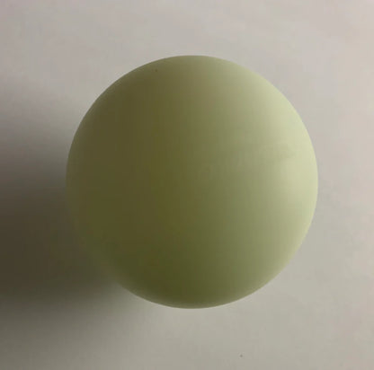 A Firefly Glow in the Dark Lacrosse Ball from the 3 pack rests on a white surface, its pale yellow-green glow casting a soft shadow left. Includes black light flashlight for charging. Brand: Firefly.