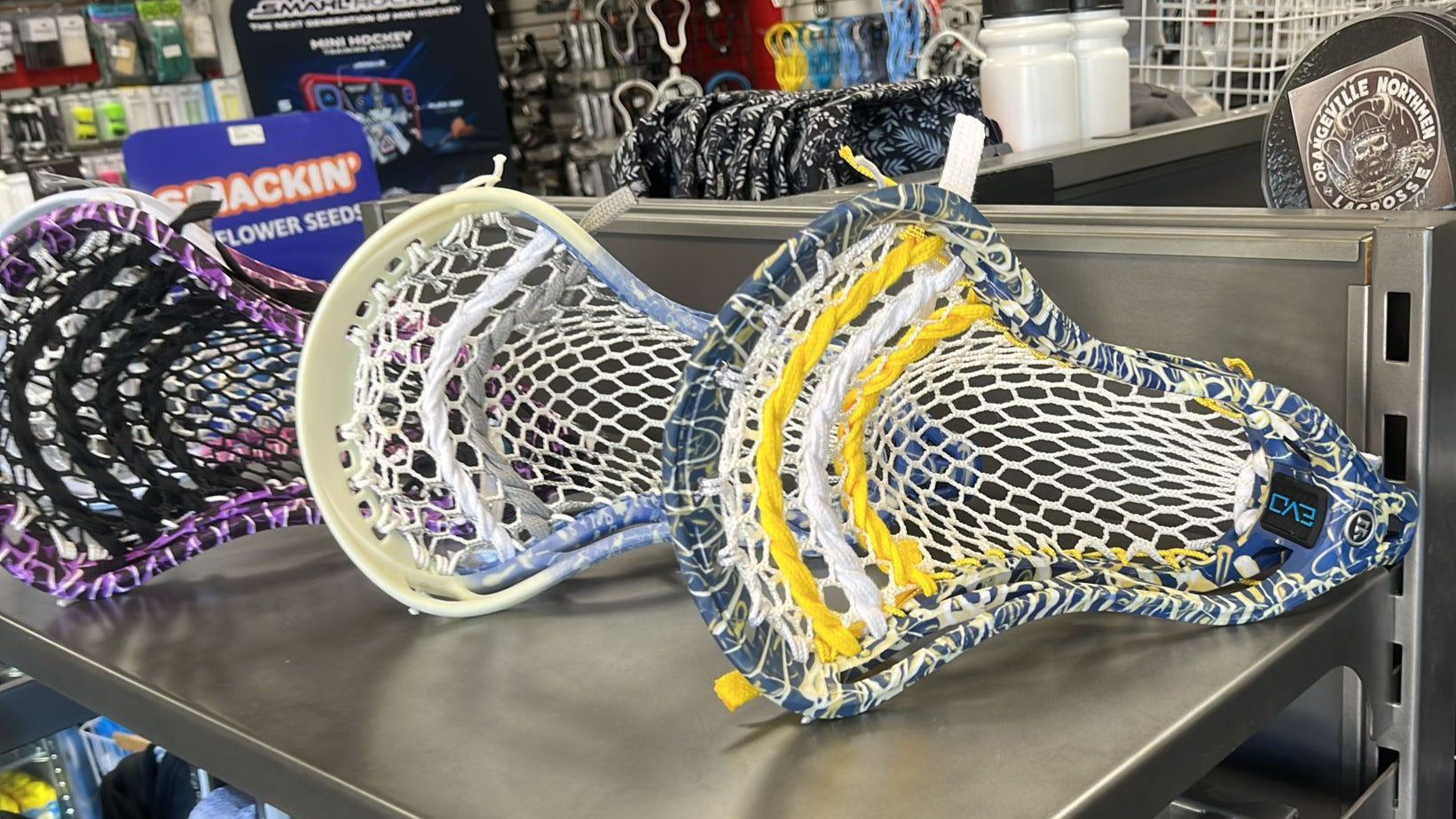 Colorful lacrosse head on a metal shelf in a store setting with various sports equipment in the background.