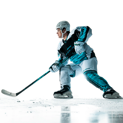 Wearing teal, black, and white, a hockey player skates on ice gripping a Bauer Pulse Hockey Stick by Bauer, eyes focused and ready for action against a bright white background.